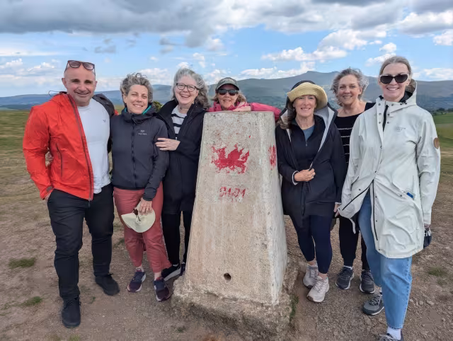John with a group of guests around a trig point painted with a red Welsh dragon, hills behind