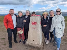 John with a group of guests around a trig point painted with a red Welsh dragon, hills behind