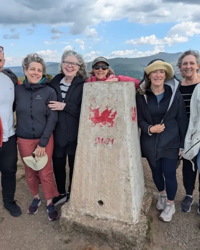 John with a group of guests around a trig point painted with a red Welsh dragon, hills behind